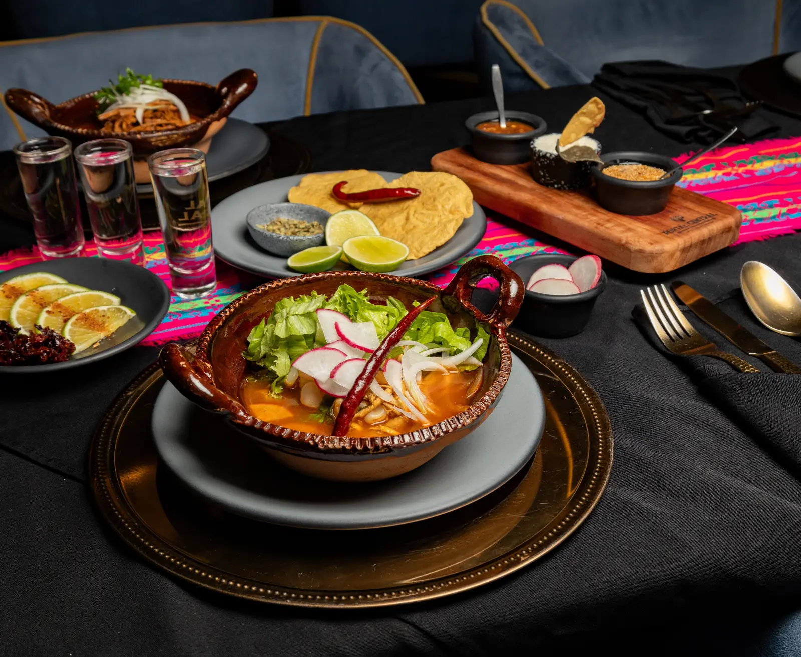 Mesa servida con pozole en cazuela de barro y camino de rebozo rosa